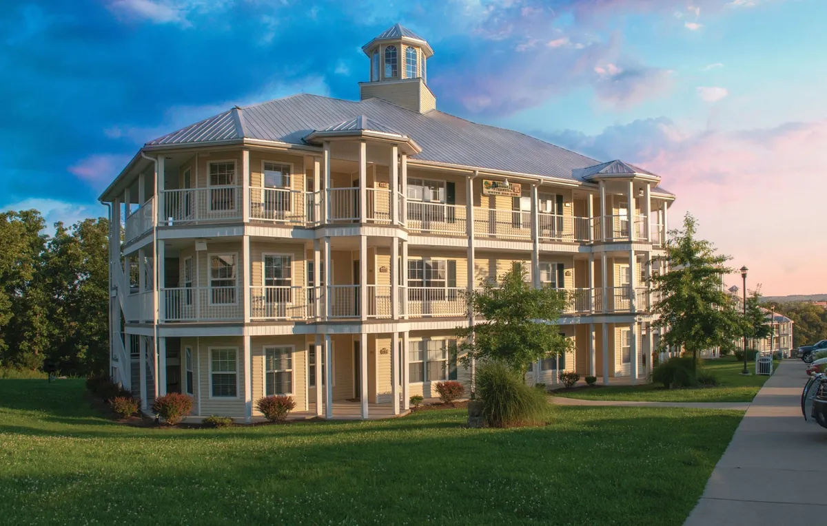 The exterior of a three-story building surrounded by a landscaped lawn, with balconies overlooking the grounds. People are seen relaxing on the balconies and strolling in the communal green spaces, creating a sense of community and leisure.