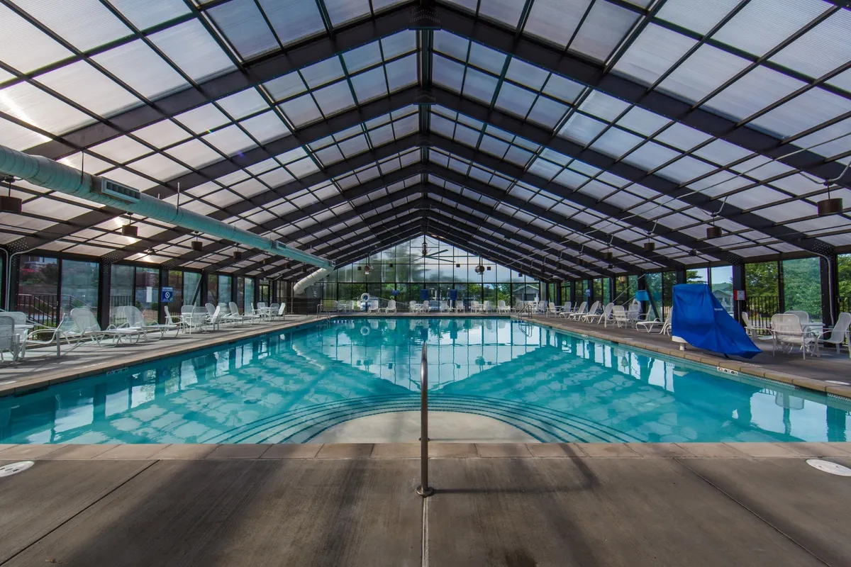 A serene indoor pool area with expansive glass ceilings letting natural light pour in. The tranquil water reflects the spacious environment, while lounge chairs line the sides, suggesting a relaxing retreat for swimming or unwinding regardless of the weather.