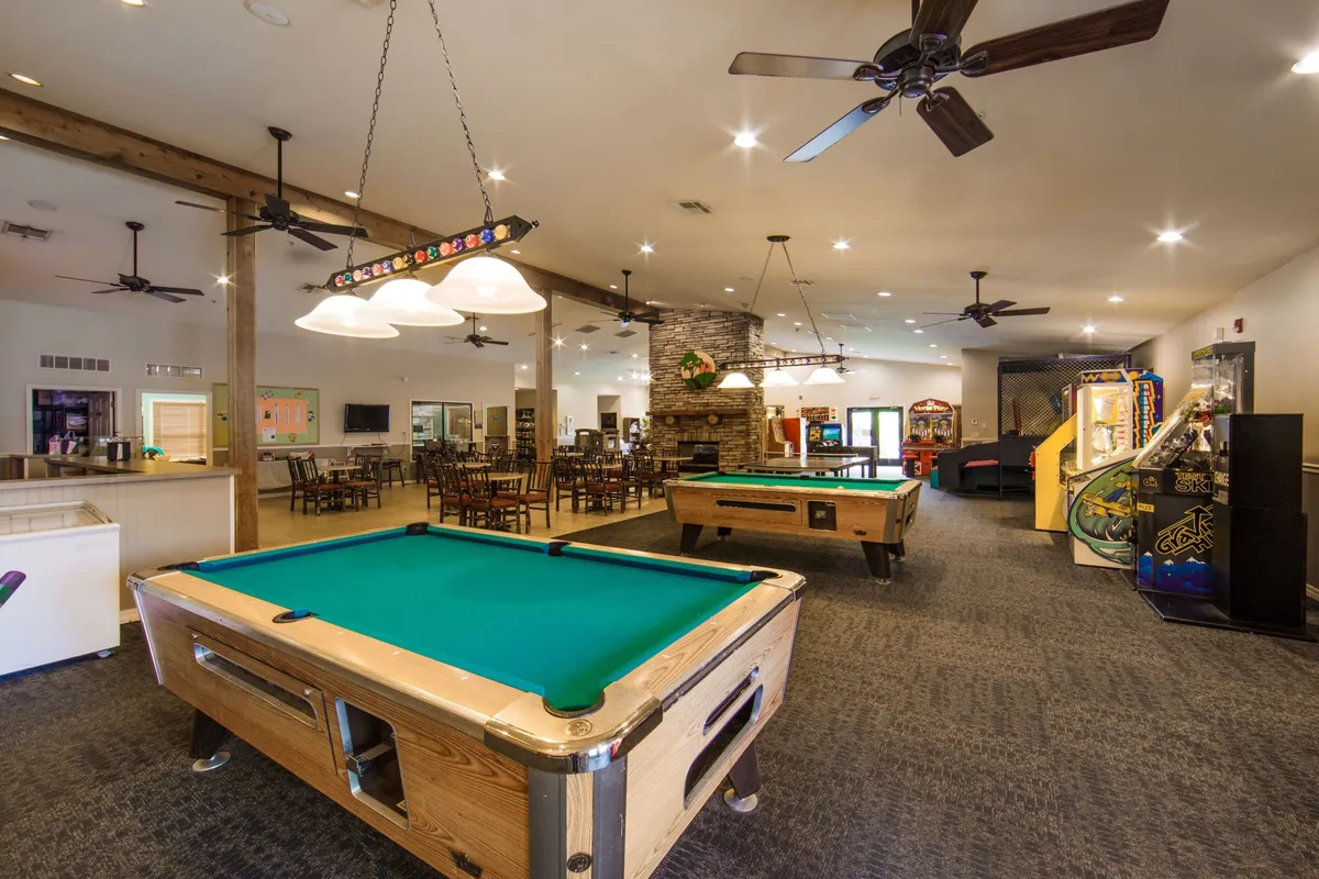 This image captures a vibrant interior scene featuring a dining area, pool tables, arcade machines, and a stone fireplace. The dining area is visible alongside spaces designed for socializing and recreation, with warm lighting and a cozy ambiance.